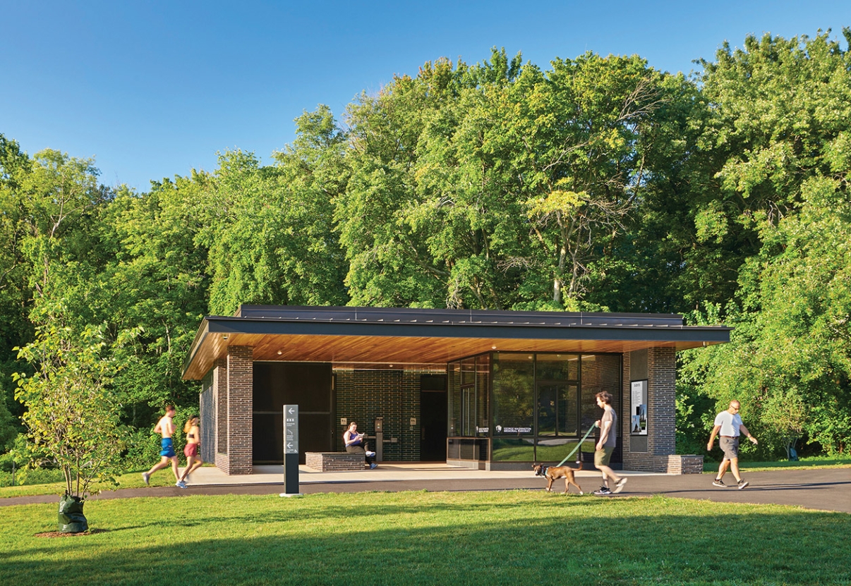 View of a small brick building across and expanse of grass. People walk and bike on baths in front of the building.