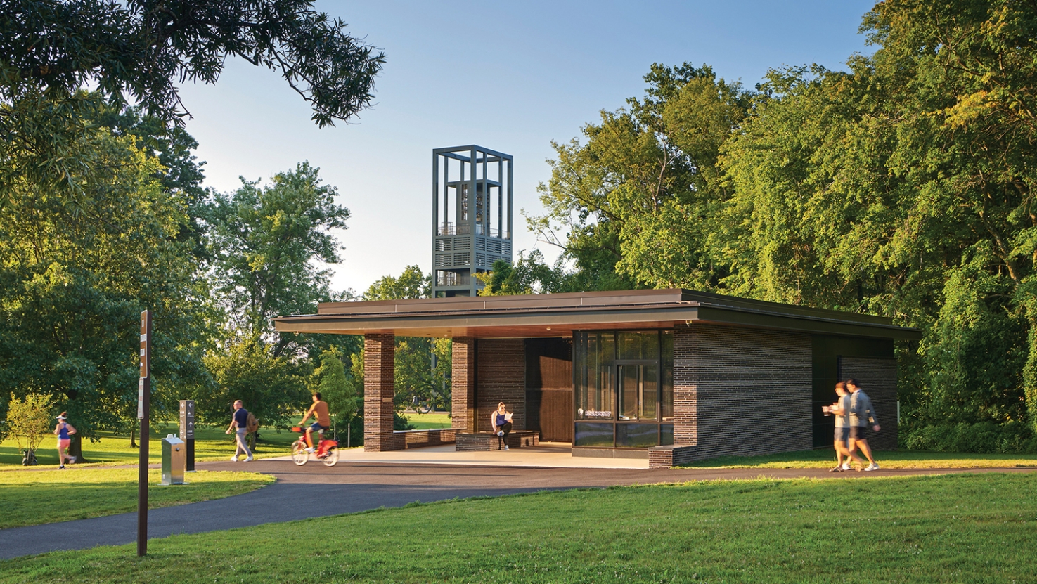 View of a small brick building across and expanse of grass. People walk and bike on baths in front of the building.