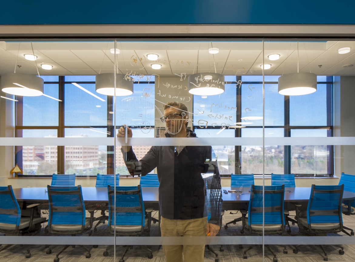 Looking through a glass wall into a conference room where a man is standing and writing notes in dry-erase marker on the glass wall.